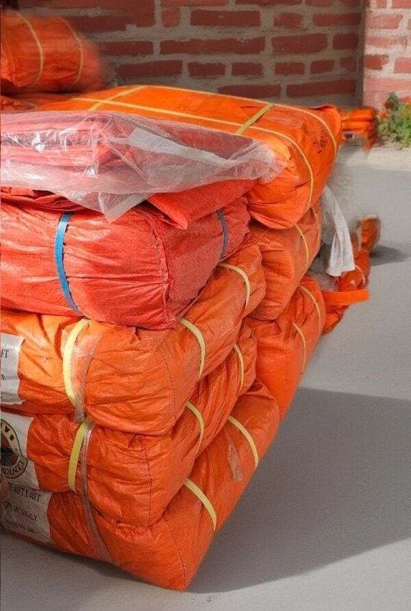 Stack of neatly bundled orange PVC Tarpaulin Sheets bags secured with straps in Abu Dhabi Store, resting on a flat surface against a brick wall.