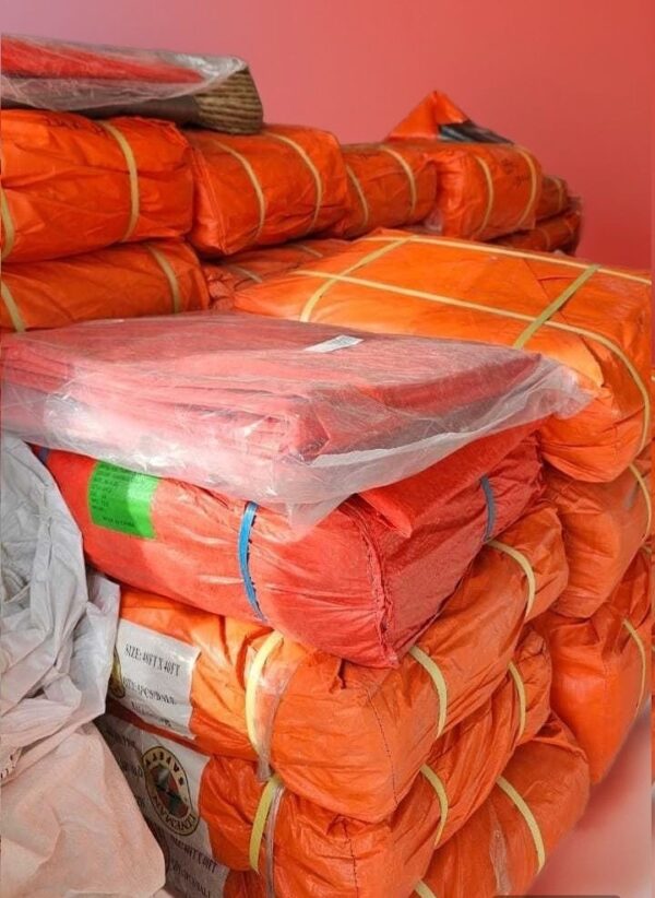 A stack of bright orange PVC Tarpaulin Sheets packages tied with bands in Abu Dhabi Store, resting against a pink wall, some partially covered with plastic wrap.